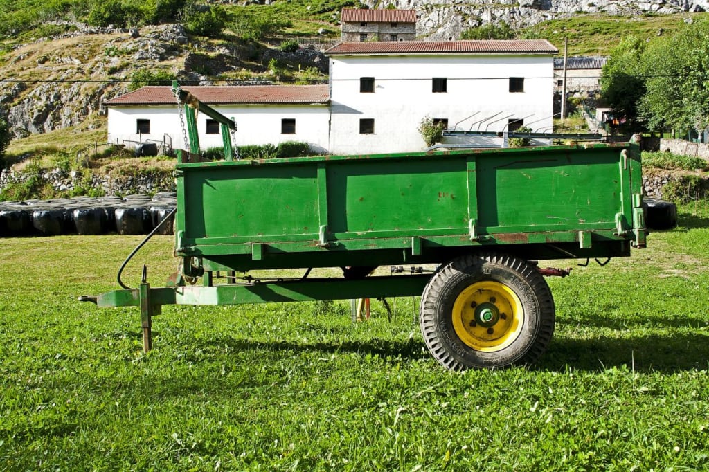 Green agricultural trailer on grass in front of rural farmhouse, used for farming or haulage